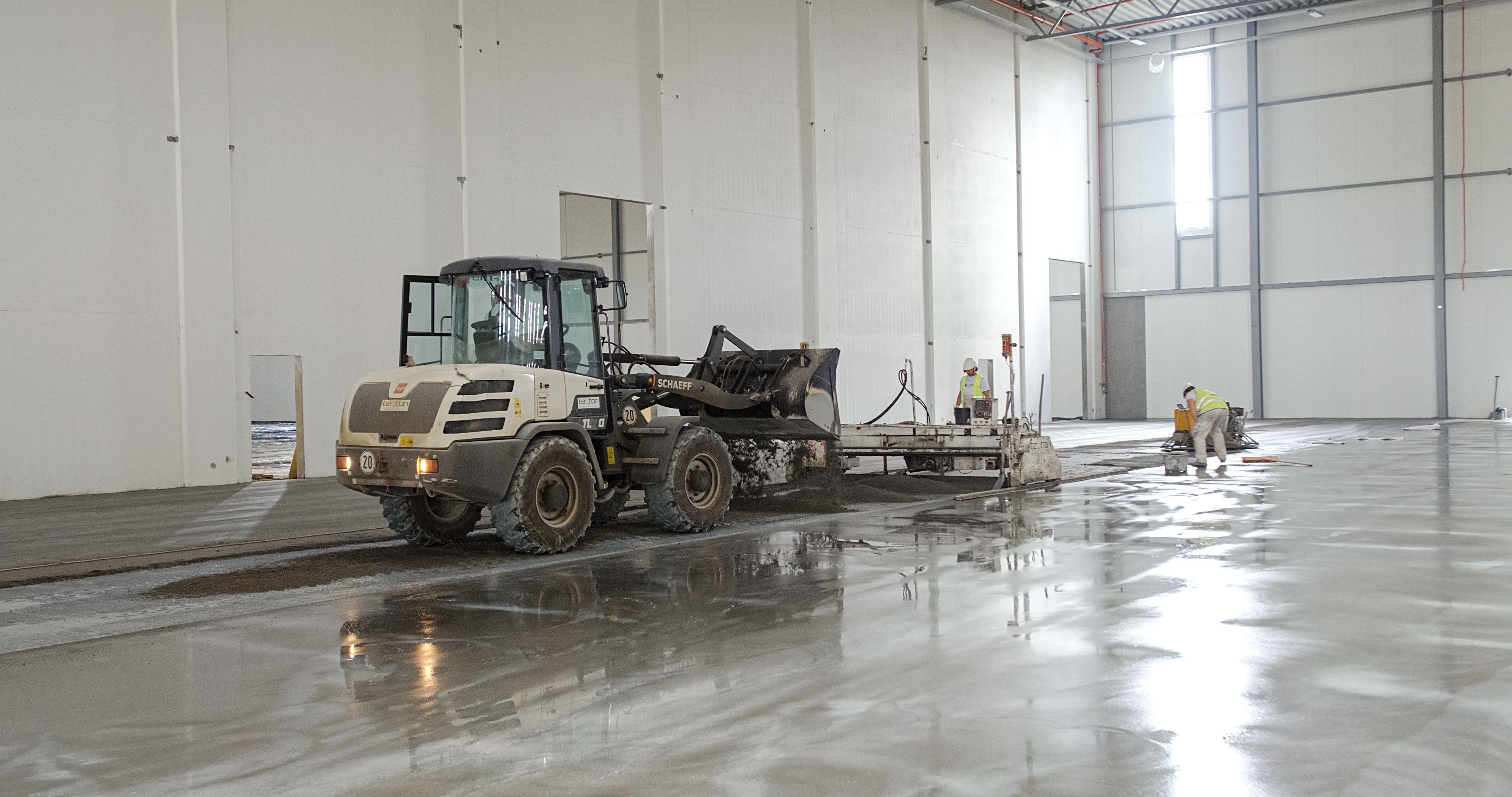 Arbetsprocess för teqton slätskikt för lagergolv med traktor samt två arbetare.