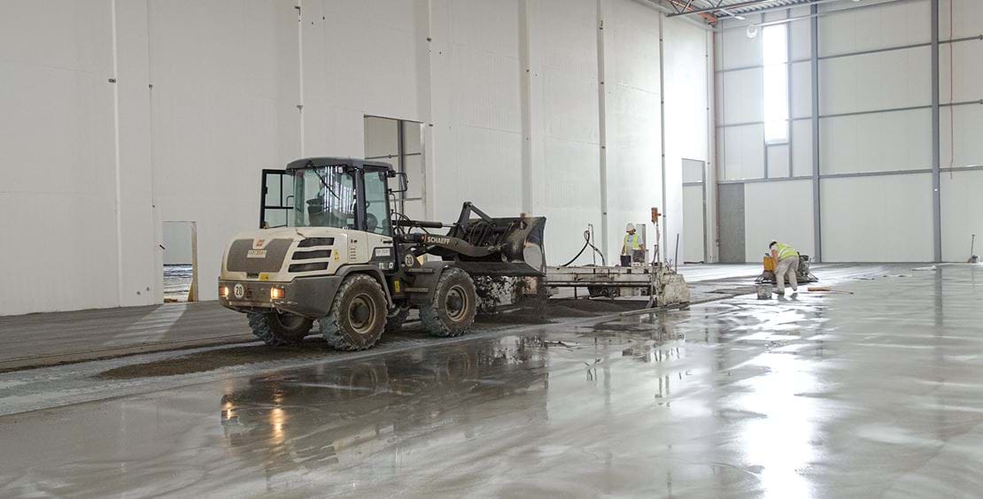 Arbetsprocess för teqton slätskikt för lagergolv med traktor samt två arbetare.